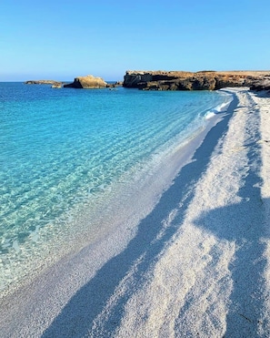 A beach with clear turquoise water, a pebbly shore, and rocky cliffs in the background.