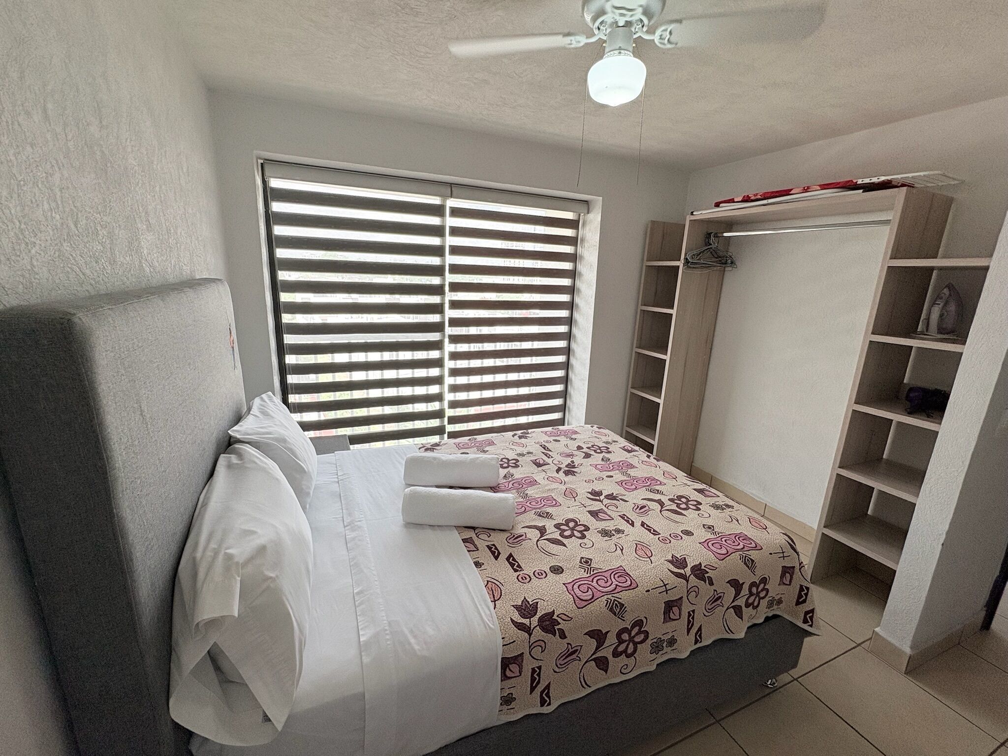 A bedroom with a bed, a ceiling fan, a window with blinds, and a built-in wardrobe.