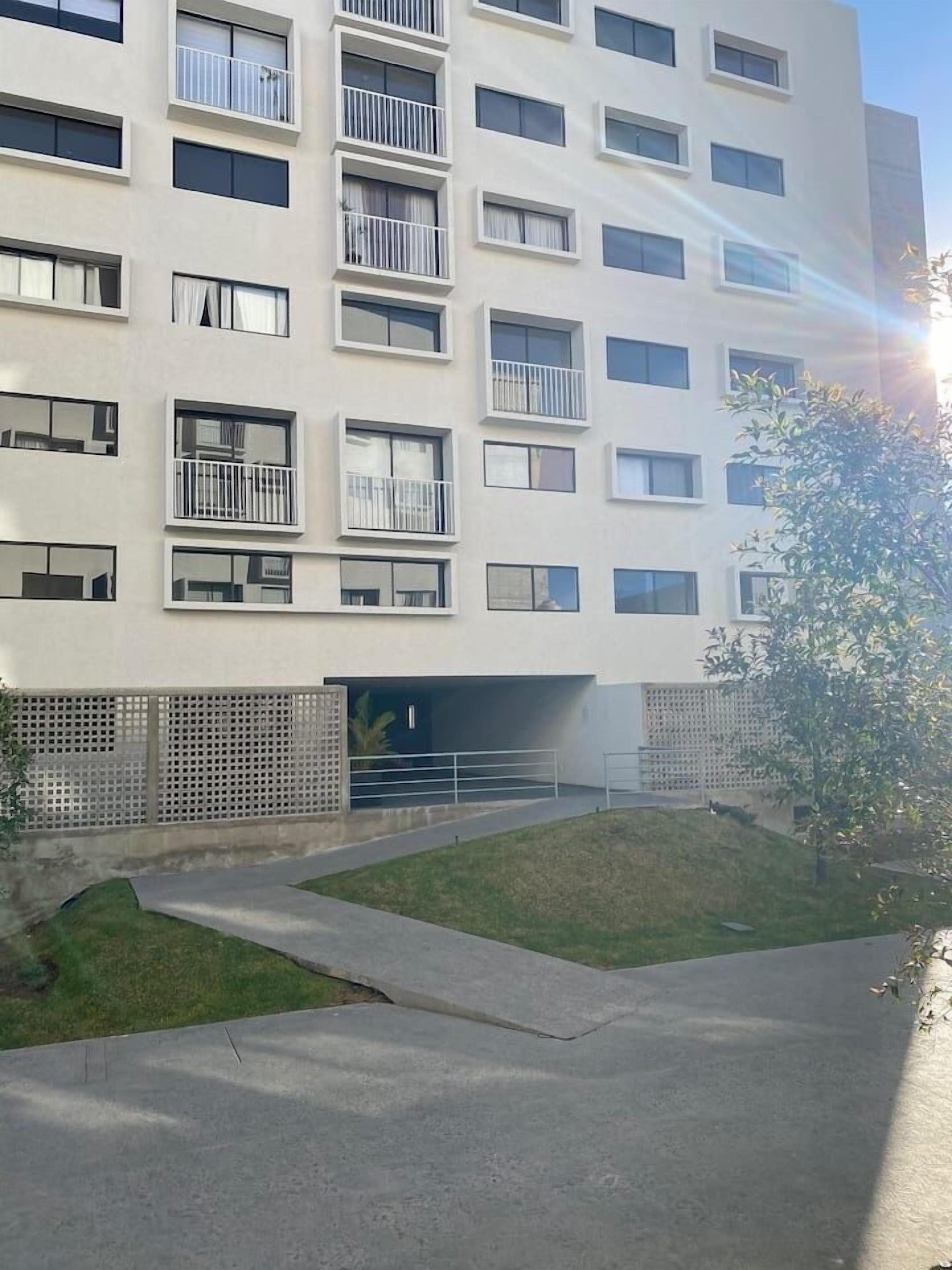 A multi-story building with balconies, a covered entrance, and a small grassy area with a pathway.