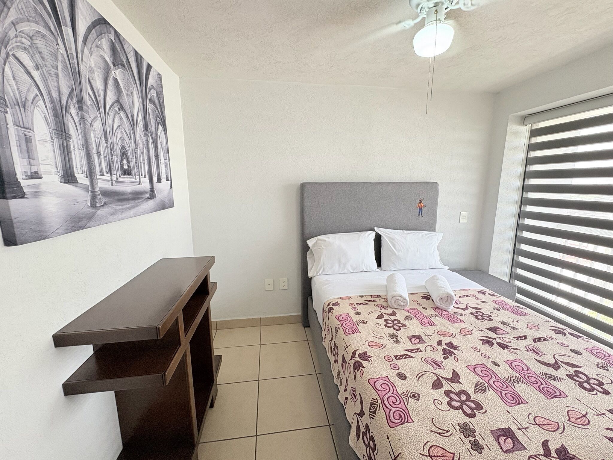 A bedroom with a bed, a nightstand, a wall-mounted shelf, a ceiling fan, and a window with blinds.