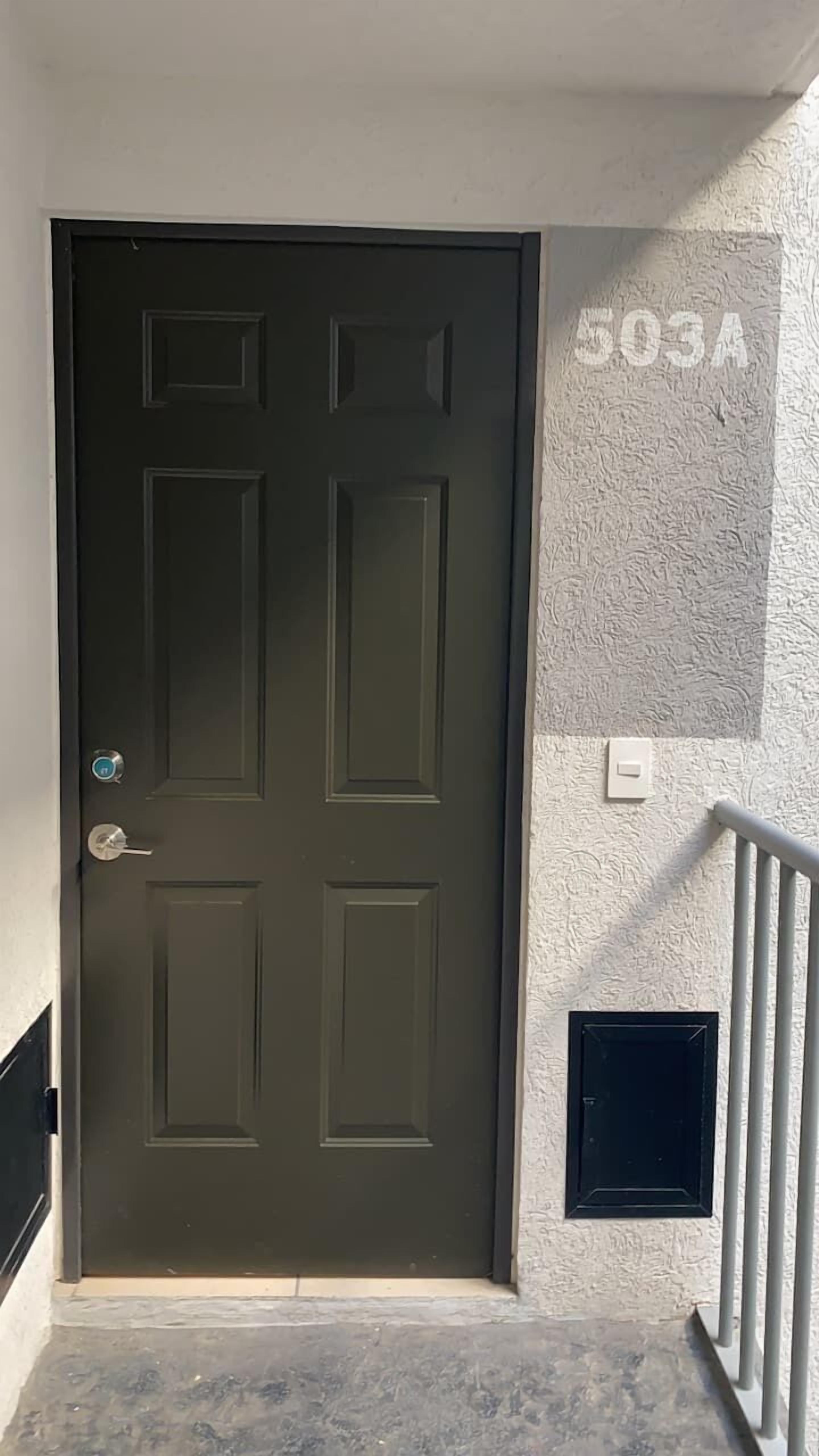 Una puerta verde oscuro con una mirilla y cerradura, un interruptor blanco pequeño, y una pared texturizada con el número '503A' pintado.