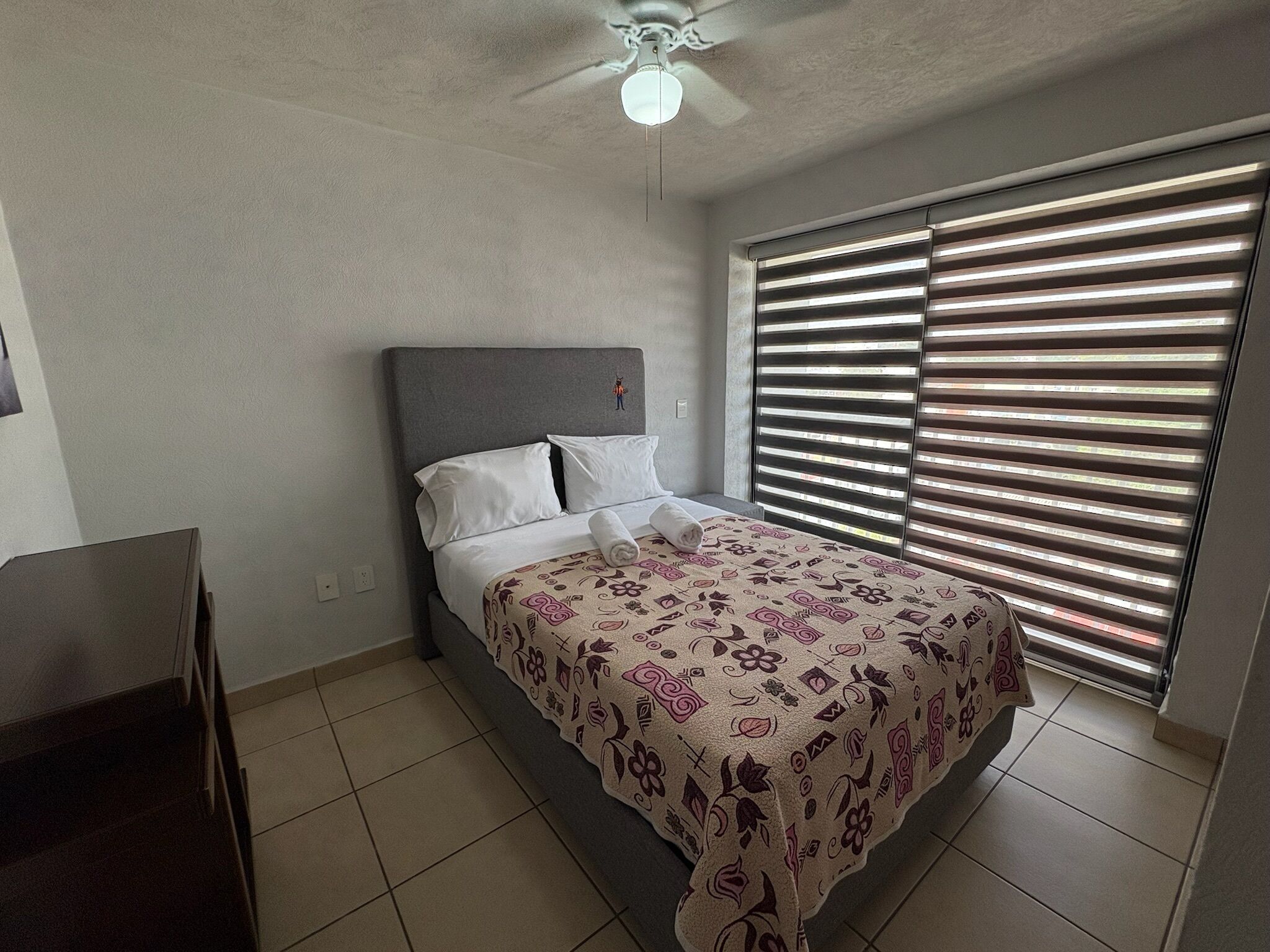 Une chambre à coucher avec un lit, un ventilateur de plafond, une fenêtre avec des stores et un meuble pour la télévision.