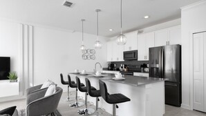 A modern kitchen with white cabinets, a central island with bar stools, stainless steel appliances, pendant lighting, and a flat-screen TV. - Modern Clean Design Near Disney Top Resort (Kissimmee)
