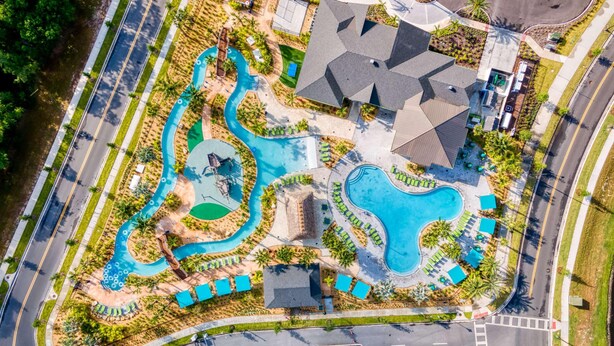 An aerial view of a resort with multiple swimming pools, a large parking area, and a central building with a sloped roof. - Modern Clean Design Near Disney Top Resort (Kissimmee)