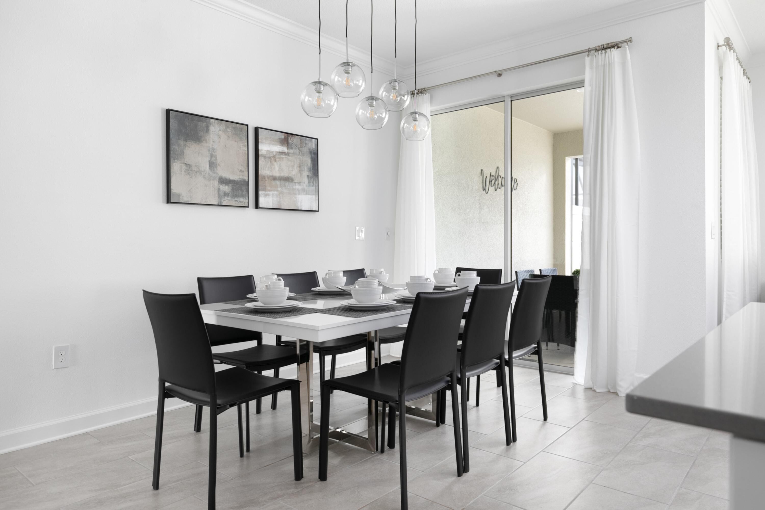 Uma sala de jantar moderna com uma mesa branca posta para seis pessoas, cadeiras pretas e iluminação pendente.