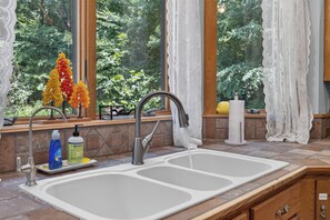 A kitchen with a double sink, a faucet, a soap dispenser, and a window with a view of trees. - Story Time Retreat (Nashville)