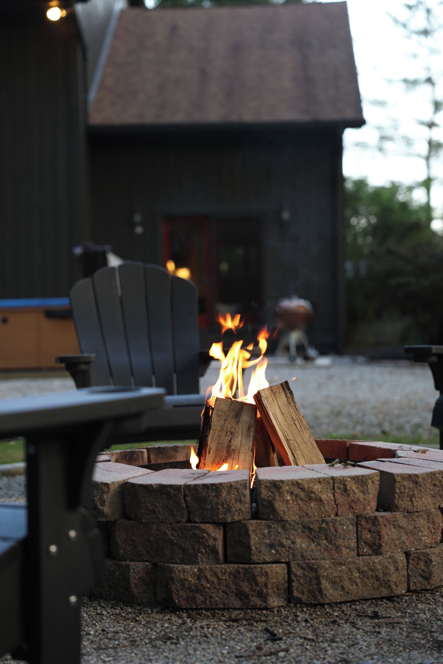 A fire pit with burning logs, a table, and chairs in a gravel area.