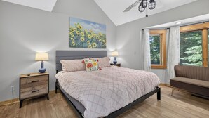 Une chambre à coucher comprenant un lit, deux tables de chevet, un ventilateur de plafond, une fenêtre avec des rideaux et un tableau accroché au mur.