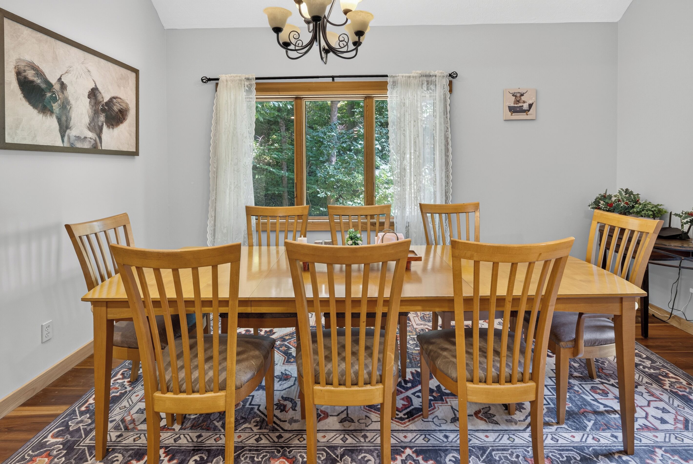 A dining room with a large table and six chairs, a chandelier, a painting on the wall, and a window with curtains.