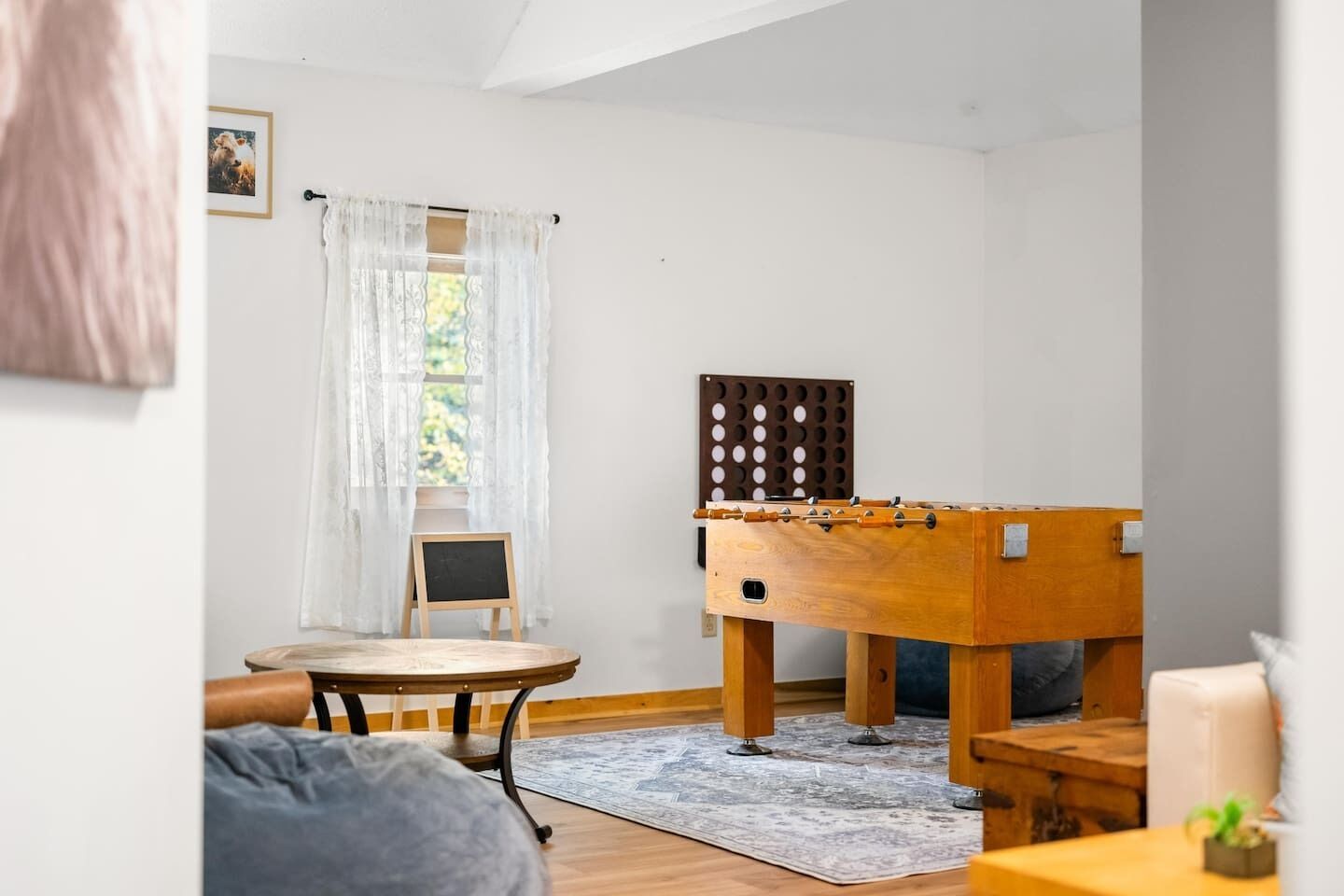 A room with a foosball table, a small wooden table, a chair, and a view of the outdoors through a window with sheer curtains.