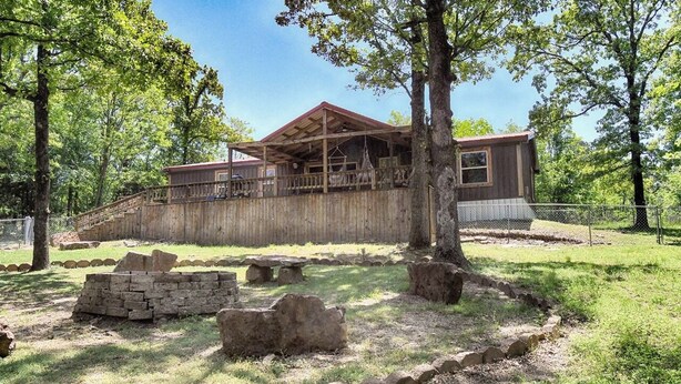 A rustic cabin with a wooden deck, surrounded by trees and stone structures.