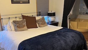 A bedroom with a bed, a nightstand, a lamp, a ceiling fan, a window with blinds, and a bathtub visible through the window.