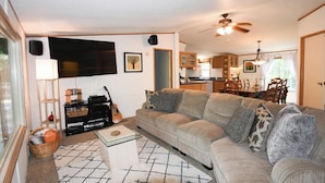 A living room with a sofa, a coffee table, a TV, and a guitar. There is a kitchen in the background.