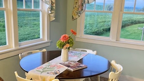 Een eetgedeelte met een ronde tafel, witte stoelen, een vaas met bloemen en uitzicht op een groen veld door ramen.