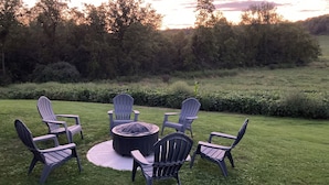 Een rond zitgedeelte buiten met Adirondack-stoelen en een vuurkorf, omringd door een verzorgd gazon en dichte bomen op de achtergrond.