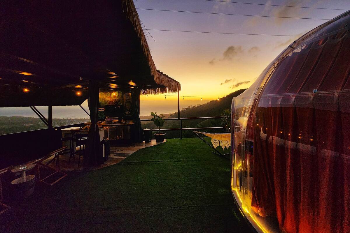 A restaurant with a view of the sunset, outdoor seating, and a thatched roof structure.