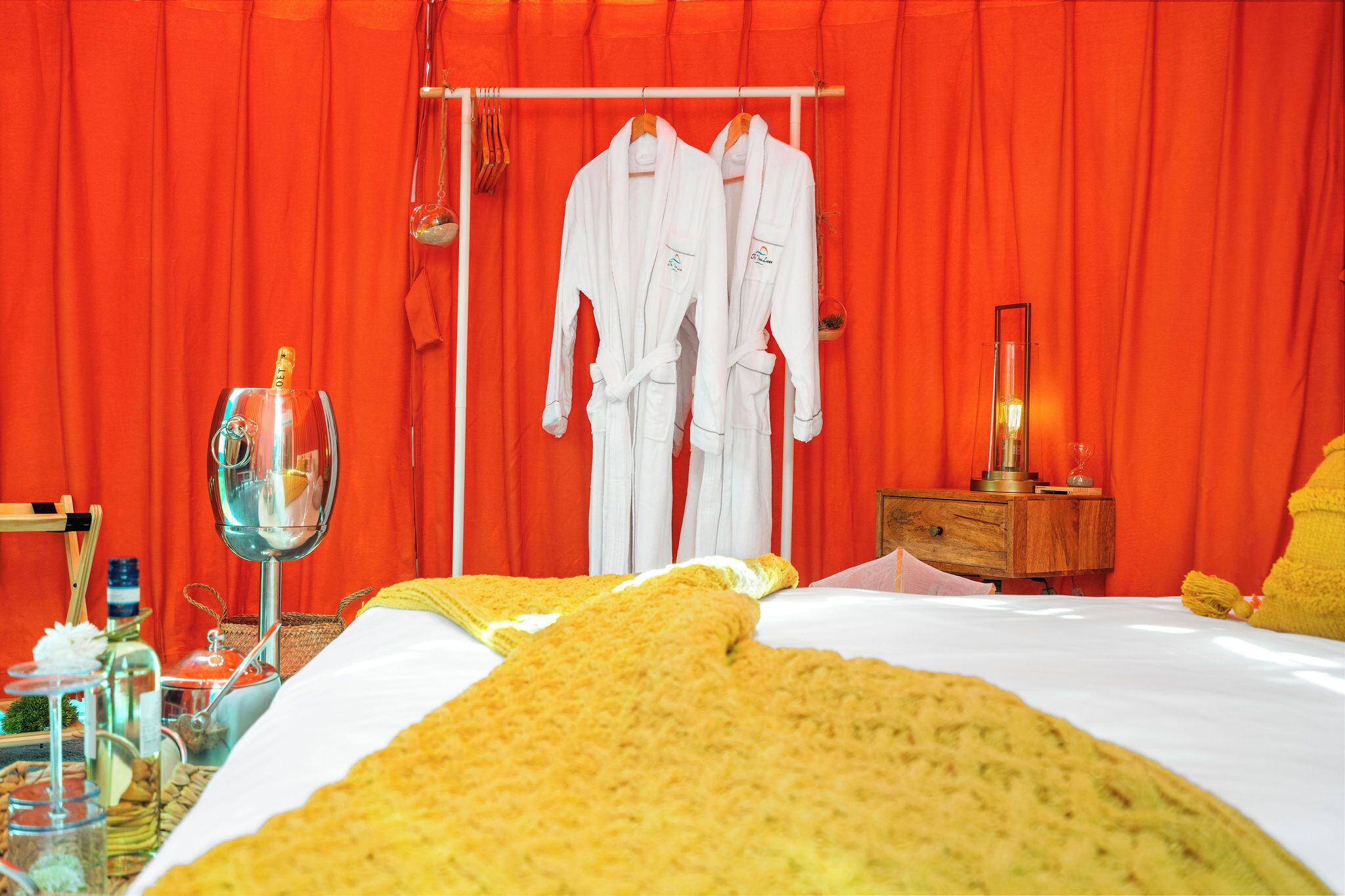 A hotel room with a bed, a bedside table, a lamp, and two white robes hanging on a rack.
