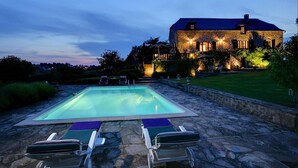 Una piscina de piedra con sillones de descanso, un sendero de piedra y un edificio de piedra con techo oscuro al fondo.