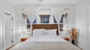Quarto com cabeceira de madeira, cama com edredom branco, duas mesas de cabeceira com abajures, janela com cortinas e uma pequena mesa de cabeceira com plume de pampa.