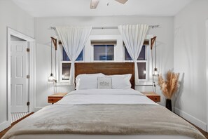 Een slaapkamer met een houten hoofdeinde, een bed met een witte deken, twee nachtkastjes met lampen, een raam met gordijnen, en een klein nachtkastje met pampa gras.