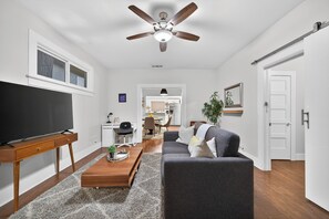 Een moderne woonkamer met een flatscreen televisie, een houten salontafel, een grijze bank, een plafondventilator en uitzicht op de keuken.