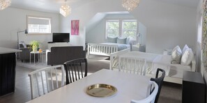A modern, well-lit room with a dining table, chairs, a white sofa, a bed with patterned bedding, a TV, and a plant. - Fieldstone Guesthouse (Clearwater)