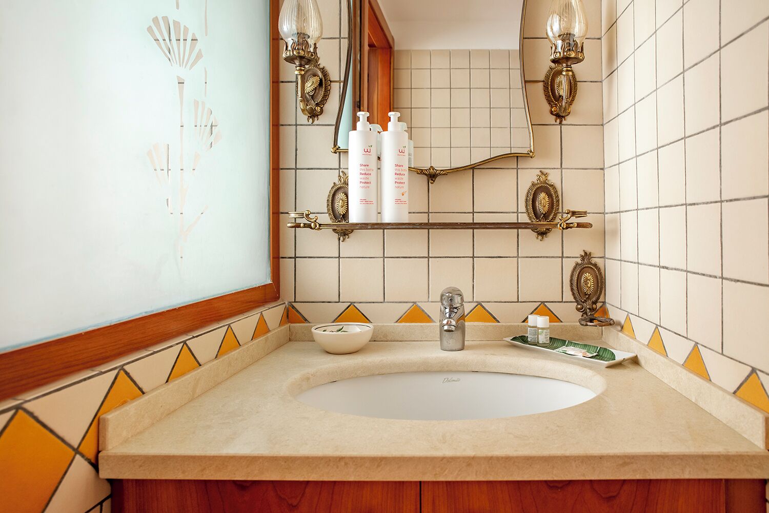 A bathroom with a large oval sink, a mirror, and two wall sconces. There are toiletries on a shelf above the sink.