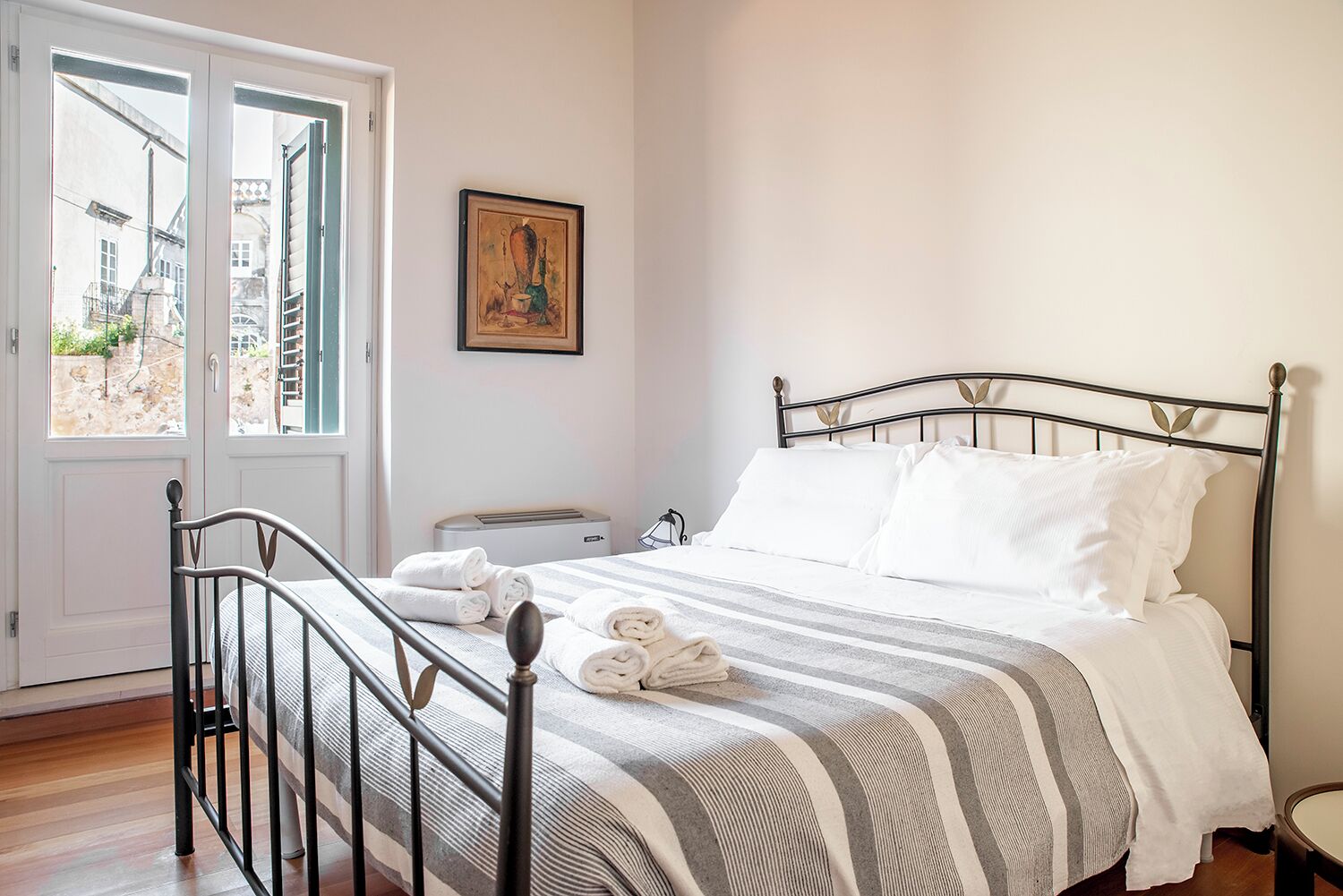 A neatly made bed with striped bedding and rolled towels, a bedside table, a radiator, and a painting on the wall.