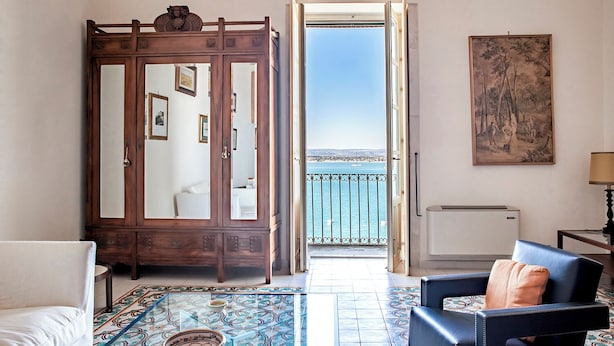 A room with a large wooden wardrobe, a view of the sea through an open door, a patterned floor, and a painting on the wall. - Duplex sul porto grande by Wonderful Italy (Syracuse)