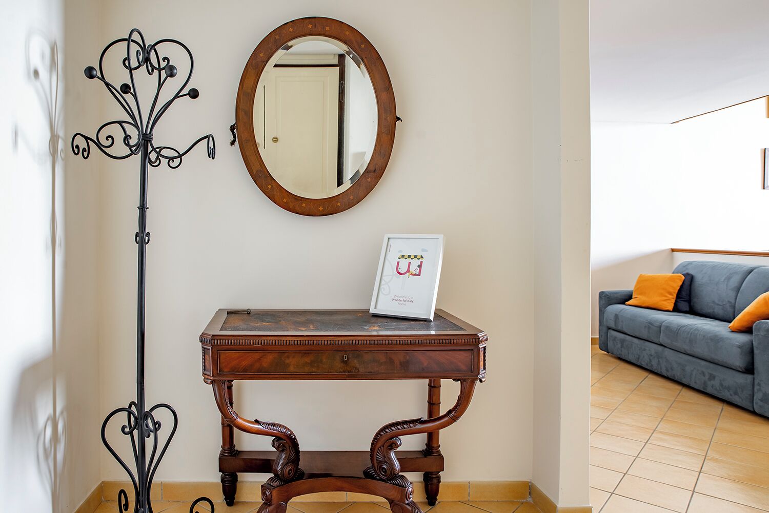 A room with a wooden console table, a mirror, a coat rack, and a blue sofa with orange cushions.