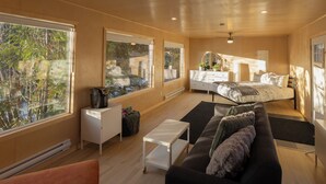 A modern bedroom with a bed, bedside table, and a dresser. There are large windows offering a view of trees and a building. - River's Edge Retreat (Clearwater)