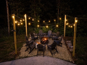 An outdoor seating area with Adirondack chairs arranged around a fire pit, surrounded by a wooden pergola with string lights.