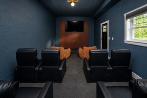 A room with a dark blue wall, a flat-screen TV mounted on a wooden panel, and a window with a view of greenery. - Modern Forest Escape w/ Pickleball, Hot Tub, Sauna, Gym, Fire Pit & Gameroom! (Mars Hill)