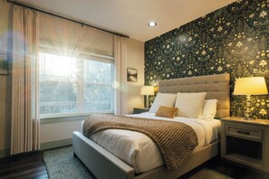 A hotel room with a large bed, two bedside lamps, a window with curtains, and a wall with a floral pattern.