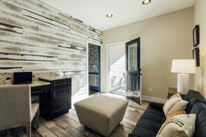 A modern living room with a wooden accent wall, a black sofa, a beige ottoman, a desk with a laptop, and a door leading to a balcony. - Modern Forest Escape w/ Pickleball, Hot Tub, Sauna, Gym, Fire Pit & Gameroom! (Mars Hill)