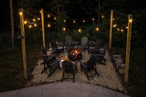 An outdoor seating area with chairs arranged around a central fire pit, surrounded by string lights. - Modern Forest Escape w/ Pickleball, Hot Tub, Sauna, Gym, Fire Pit & Gameroom! (Mars Hill)