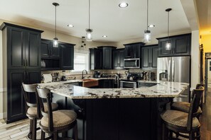 A kitchen with dark cabinets, a large island with bar stools, stainless steel appliances, and pendant lighting. - Modern Forest Escape w/ Pickleball, Hot Tub, Sauna, Gym, Fire Pit & Gameroom! (Mars Hill)
