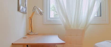 A room with a wooden desk, a white chair, a lamp, and a window with sheer curtains.