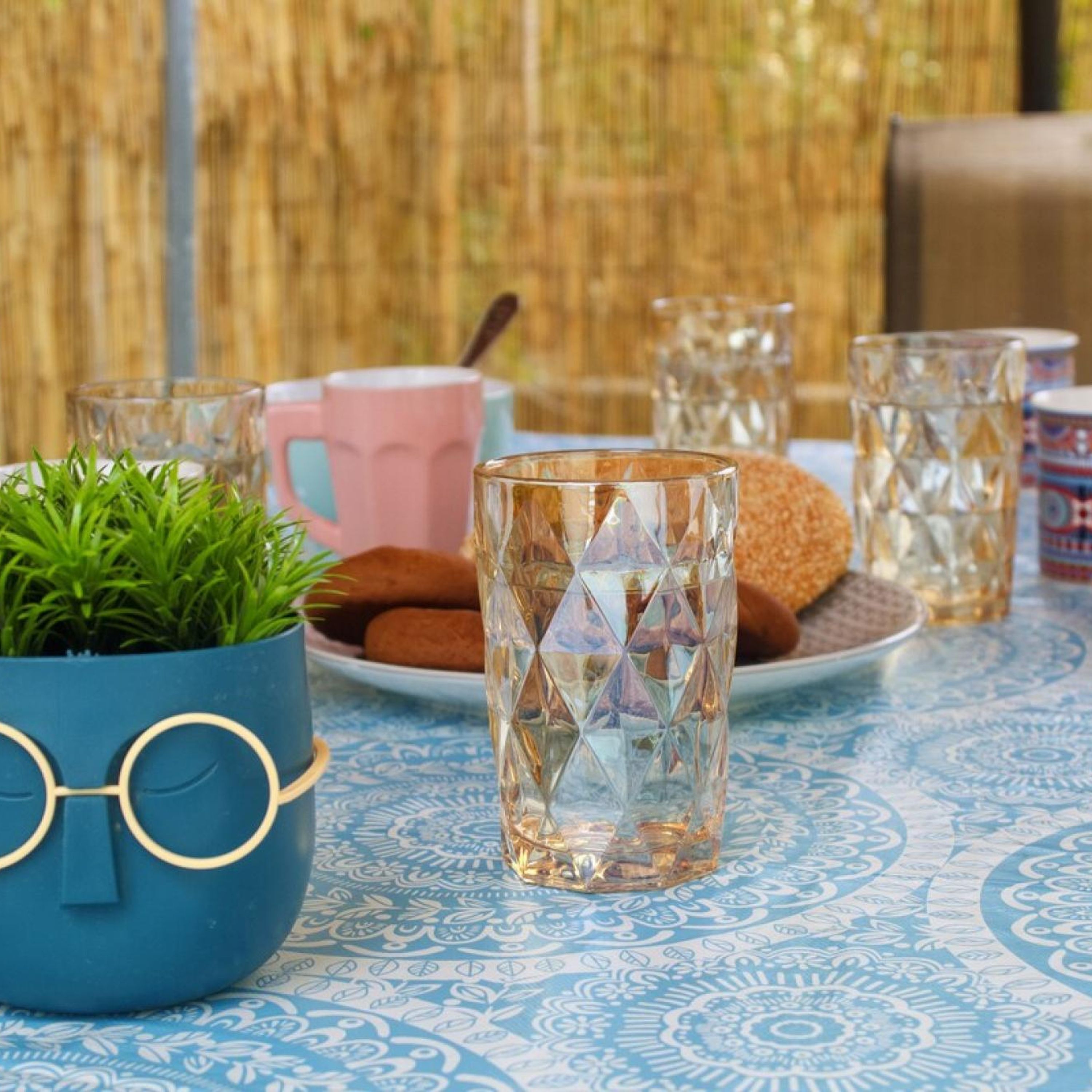Ein Tisch mit einer gemusterten Tischdecke, ein blauer Topf mit einer Pflanze, ein Teller mit Keksen und mehrere Gläser.