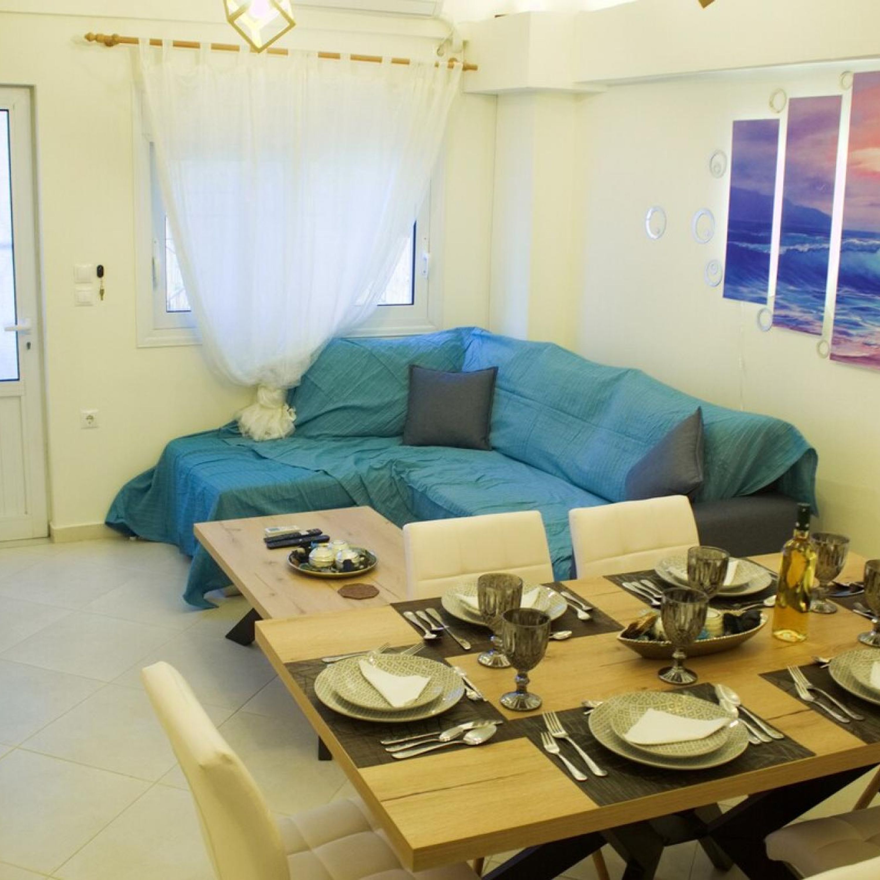 Un salon chaleureux avec un canapé bleu, une table à manger dressée pour quatre personnes et une vue sur la mer par la fenêtre.