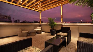 A rooftop terrace with wicker furniture, a potted plant, and a view of the city skyline at dusk.