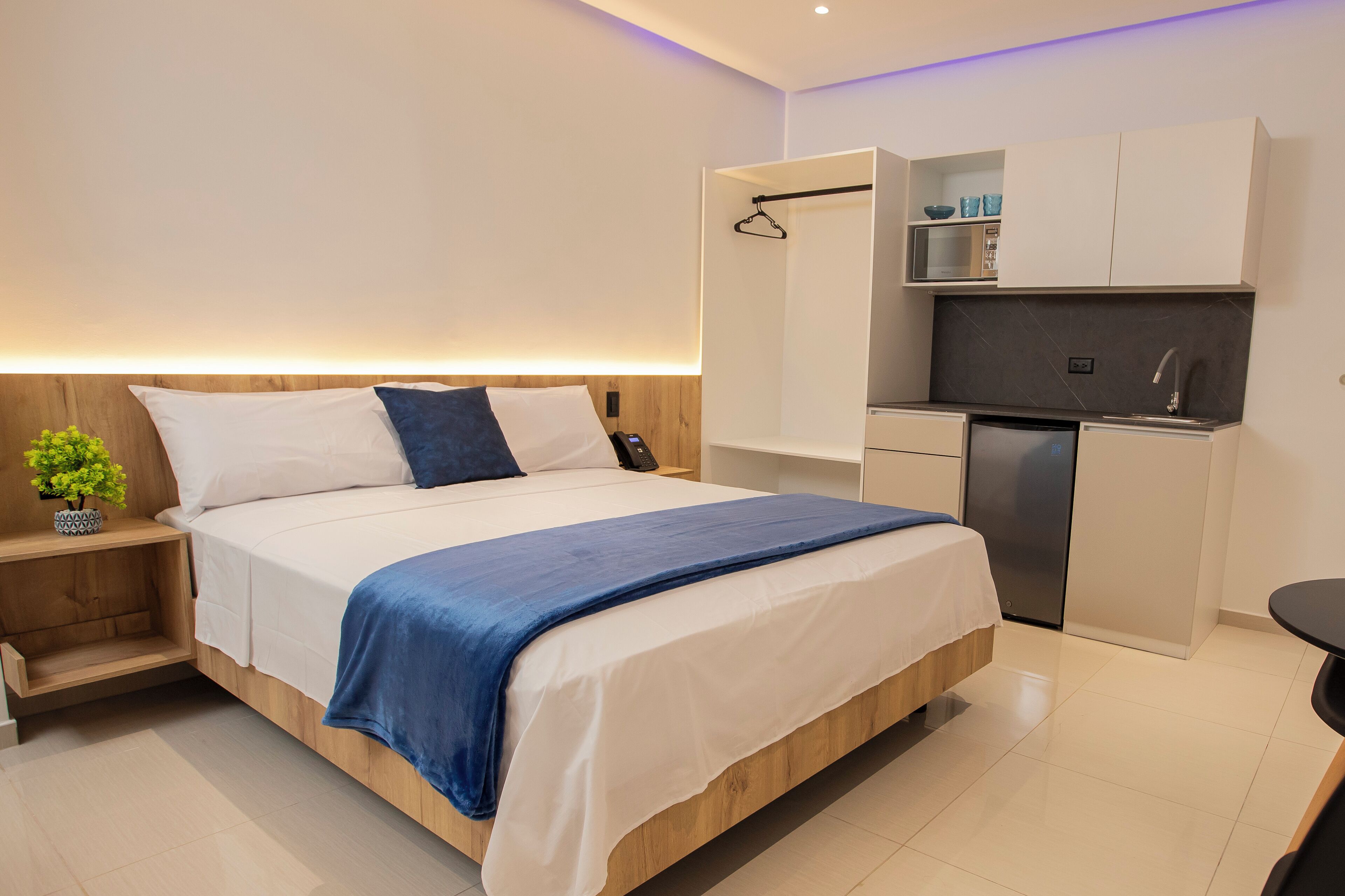Una habitación de hotel moderna con una cama de madera, una mesita de noche pequeña con una planta, una zona de cocina integrada con microondas y refrigerador, y un perchero.
