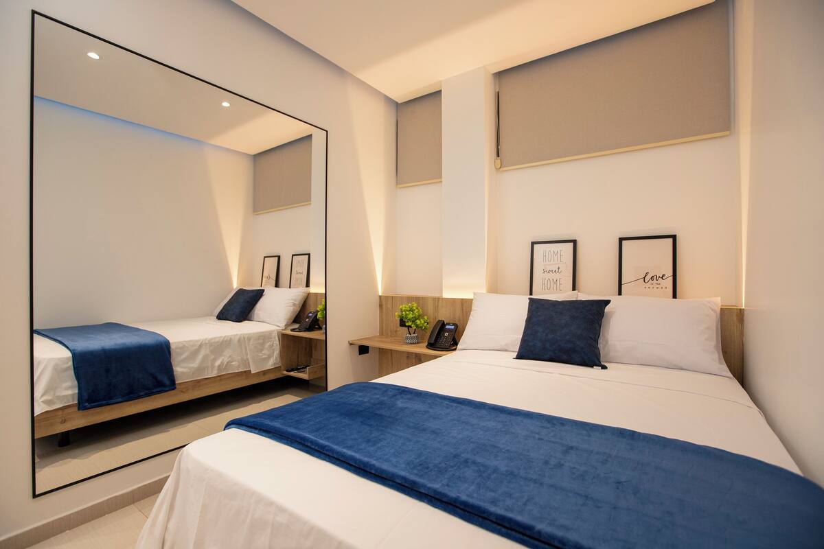 Ein modernes Hotelzimmer mit zwei Betten, einem großen Spiegel, Nachttischen mit Lampen und gerahmten Bildern an der Wand.