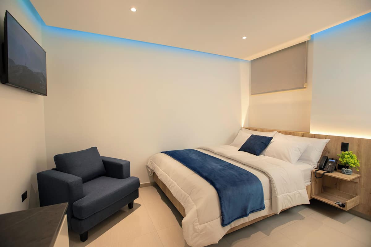 Ein modernes Schlafzimmer mit einem Bett, einem Sessel, einem Nachttisch mit Telefon und einem an der Wand befestigten Fernseher.