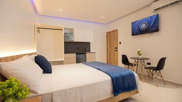 A modern hotel room with a bed, a small kitchenette, a dining area with a table and chairs, and a TV mounted on the wall.