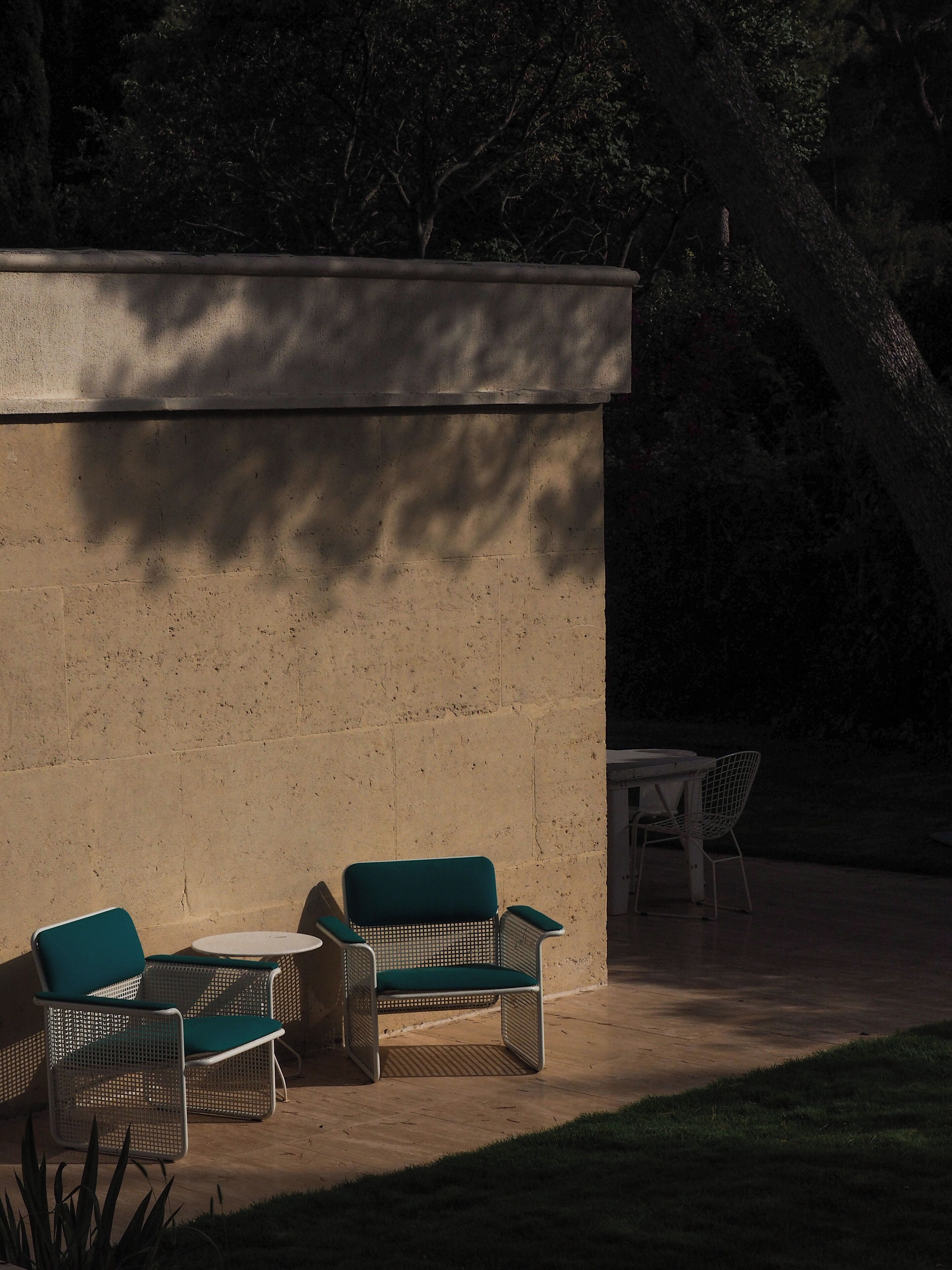 Un área de estar al aire libre con mobiliario de mimbre y una mesa pequeña, frente a un muro de piedra con sombras proyectadas por árboles cercanos.