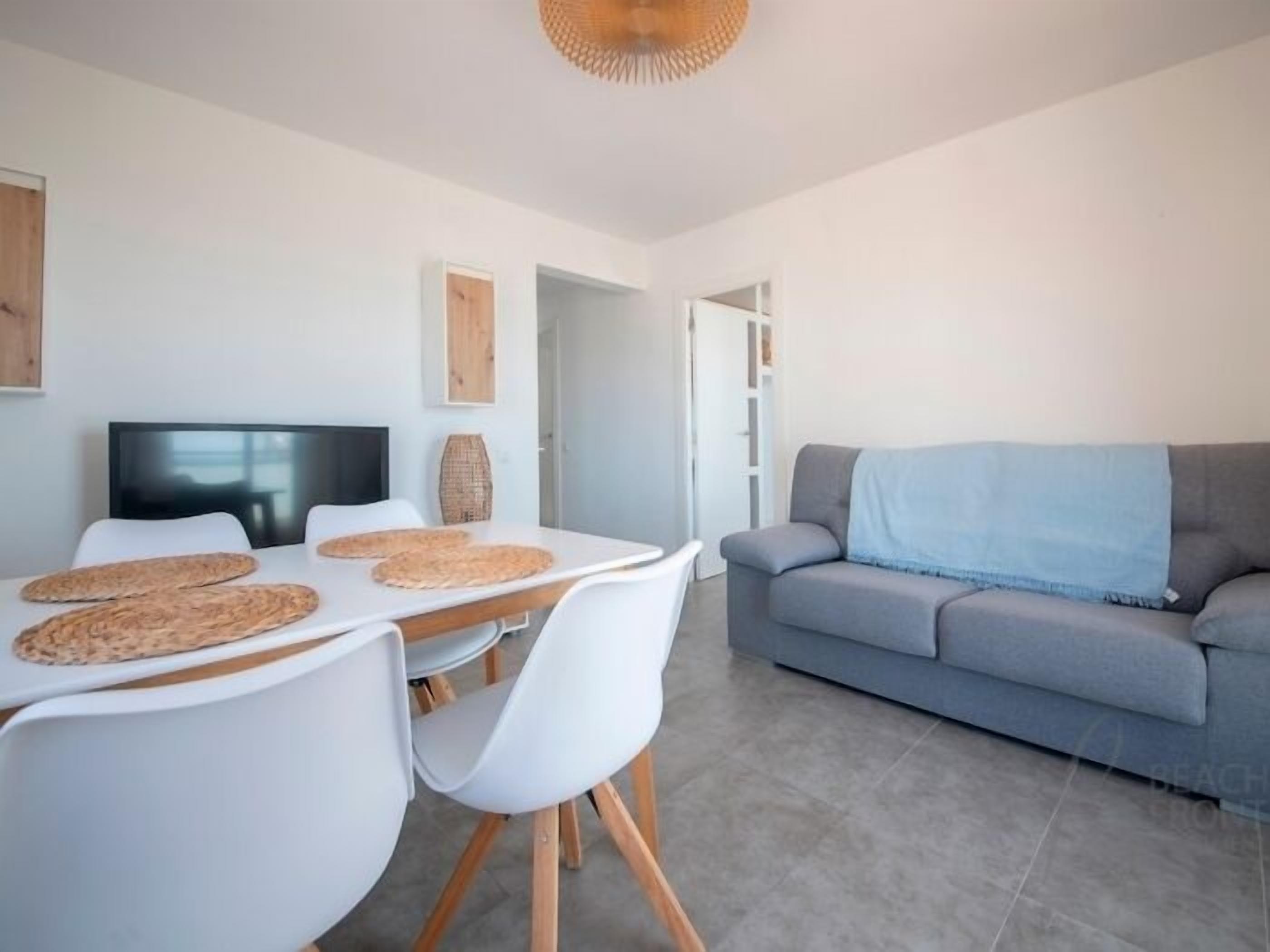 Een moderne woonkamer met een grijze bank, witte stoelen, een eettafel met placemats, een flatscreen televisie en een geweven mand aan de muur.
