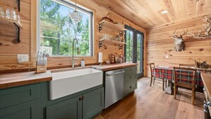 A wooden cabin interior with a kitchen featuring a large window, wooden shelves, and a deer head decoration.