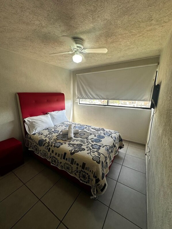 A bedroom with a bed, a red headboard, a ceiling fan, a window with blinds, and a red bench.