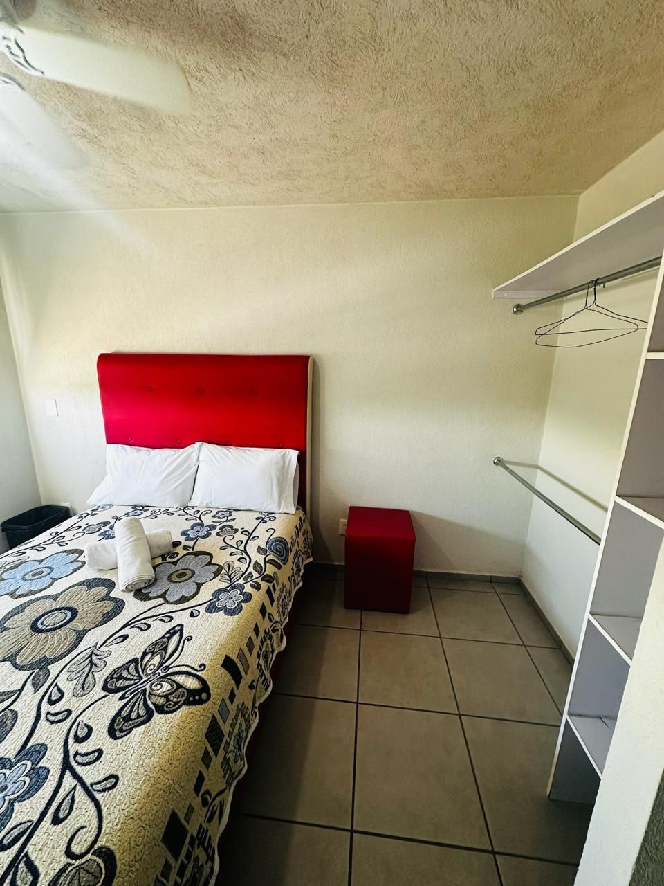 A hotel room with a bed, a red headboard, a small red nightstand, and a white wardrobe with hanging space.